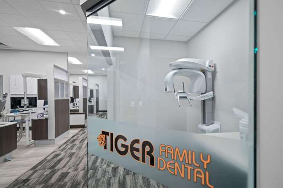 The image is a photograph of an interior space, specifically a dental office, featuring a modern reception area with a glass wall displaying the name TIGER FAMILY DENTAL and a logo, along with a digital sign-in kiosk. The room has a contemporary design with light wood flooring, neutral tones, and professional furnishings.