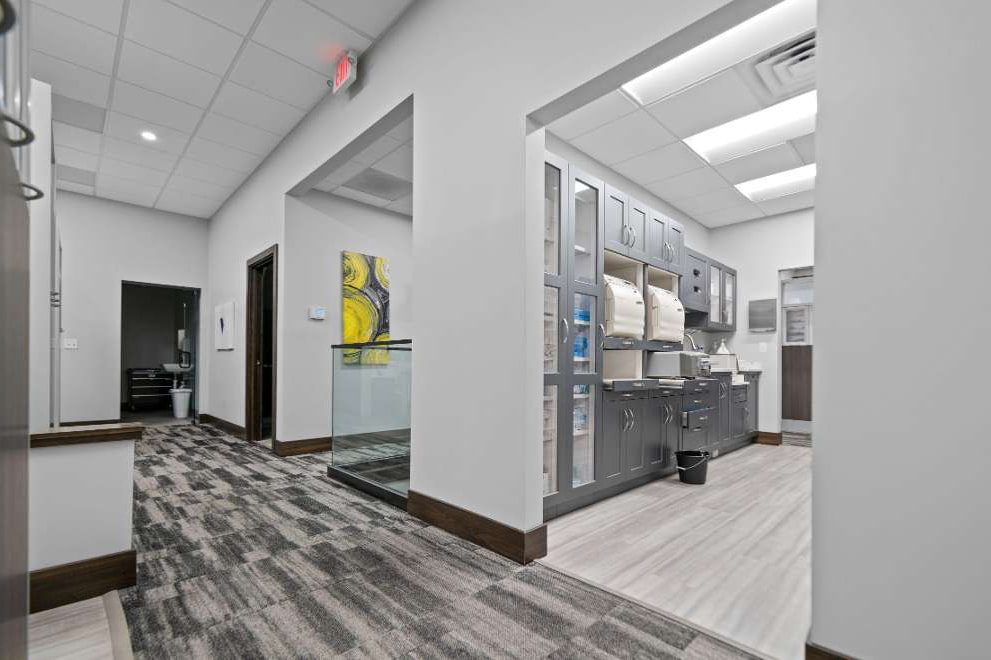 The image shows an interior space that appears to be a modern office or professional environment with a well-lit hallway leading to a room, featuring a reception area with a desk and chairs, and a large window allowing natural light into the space.