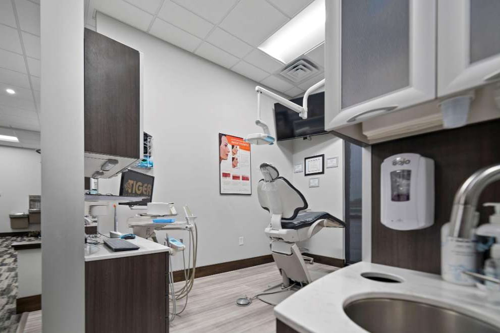 The image shows an interior view of a dental office with a dental chair, sink, and various equipment typical of such a setting.
