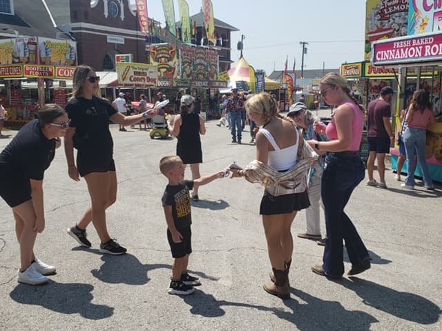 The image depicts a bustling carnival scene with people standing around a food stand, enjoying their time at the fair.
