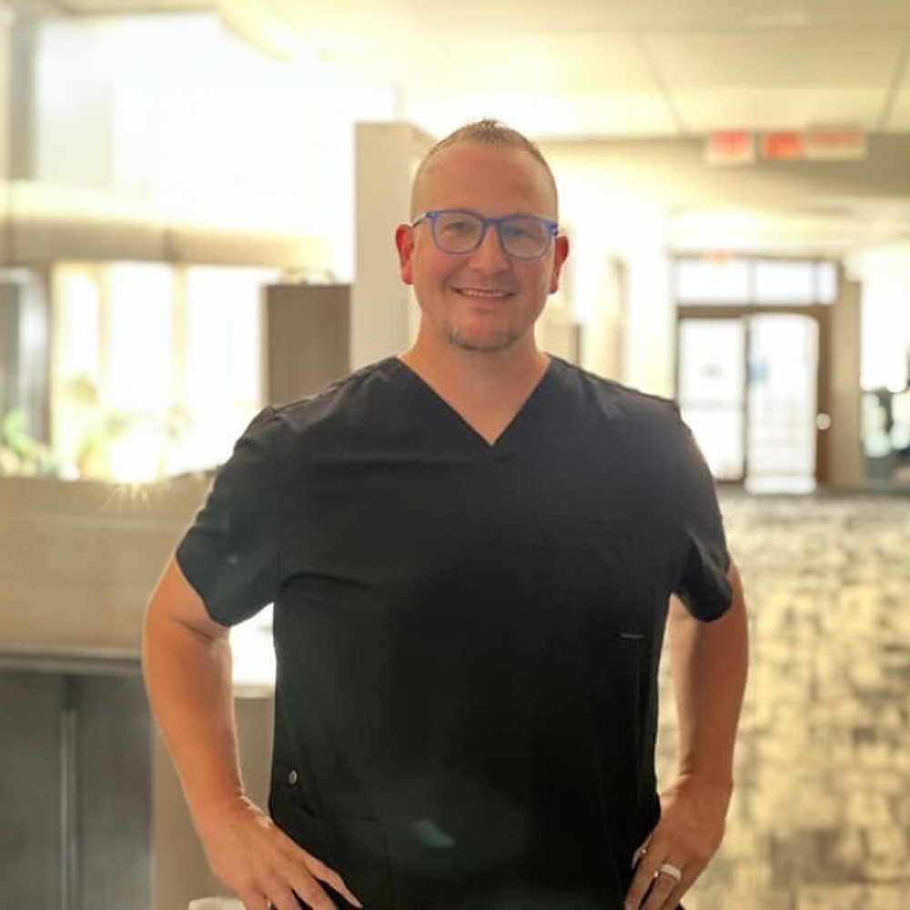 The image features a man wearing glasses, standing inside an office building with a professional appearance, posing for a portrait-style photograph.