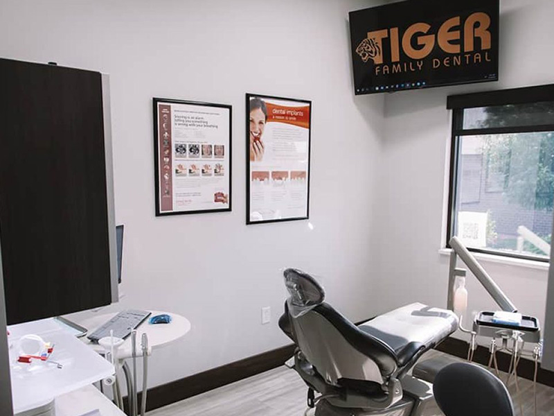 The image shows an interior view of a dental office reception area with a sign for Tiger Family Dental on the wall, featuring a seating area with pamphlets and a computer screen displaying information.
