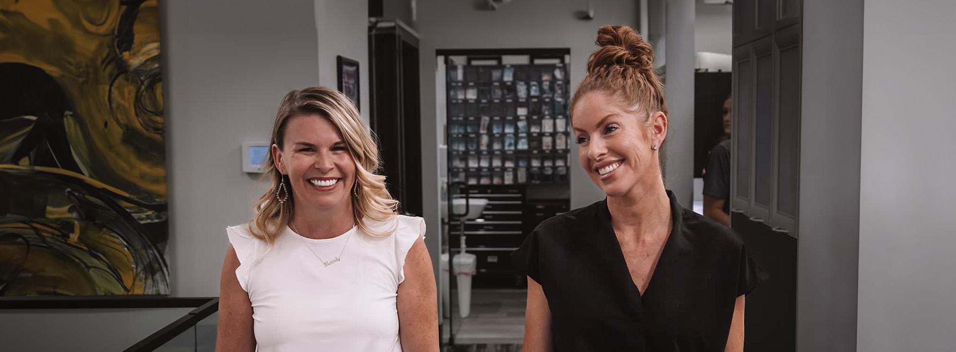 The image shows two individuals standing side by side with one person slightly ahead of the other, both smiling at the camera. They are indoors, possibly a professional setting given the presence of office-style furniture in the background.