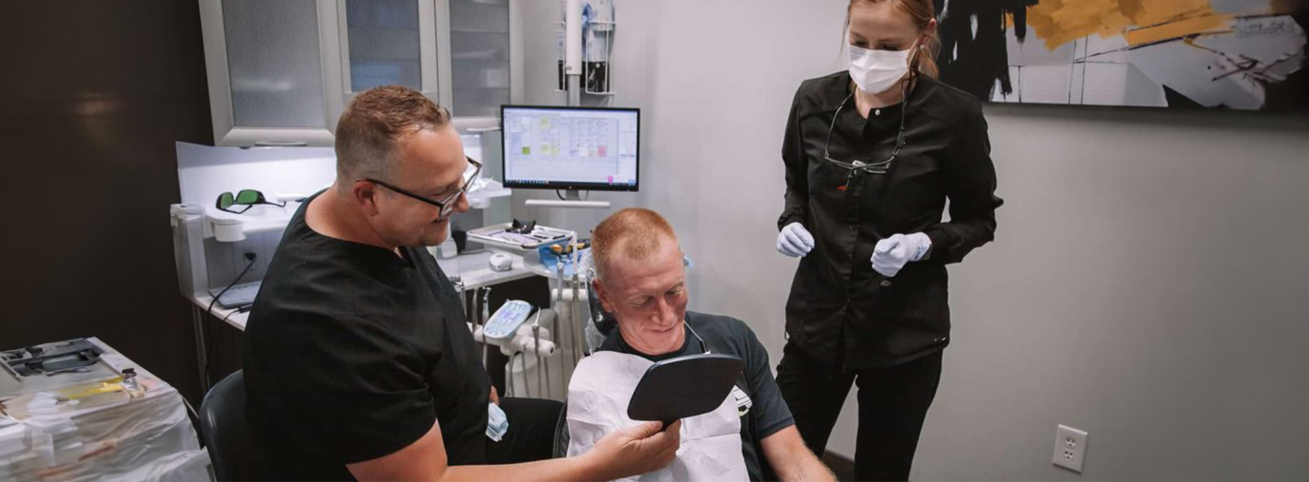 The image shows a group of people gathered around a man seated in a chair with his head tilted back, wearing a medical mask, and holding a tablet computer.