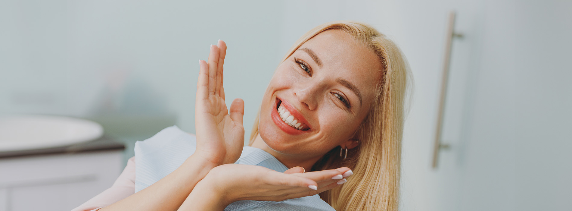 The image shows a woman with her mouth open, smiling broadly while making hand gestures, against a blurred background that suggests an indoor setting with a blue-green hue.