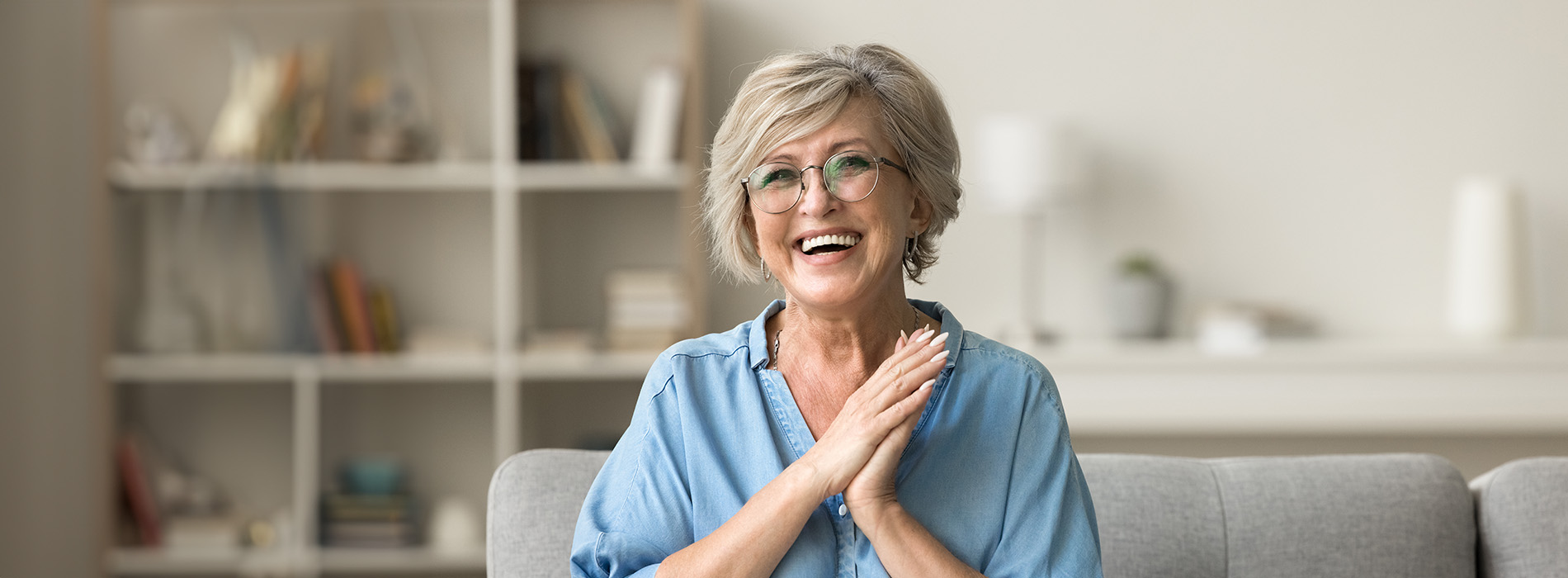 The image shows an older woman with her hands clasped together, smiling at the camera, sitting on a couch indoors.