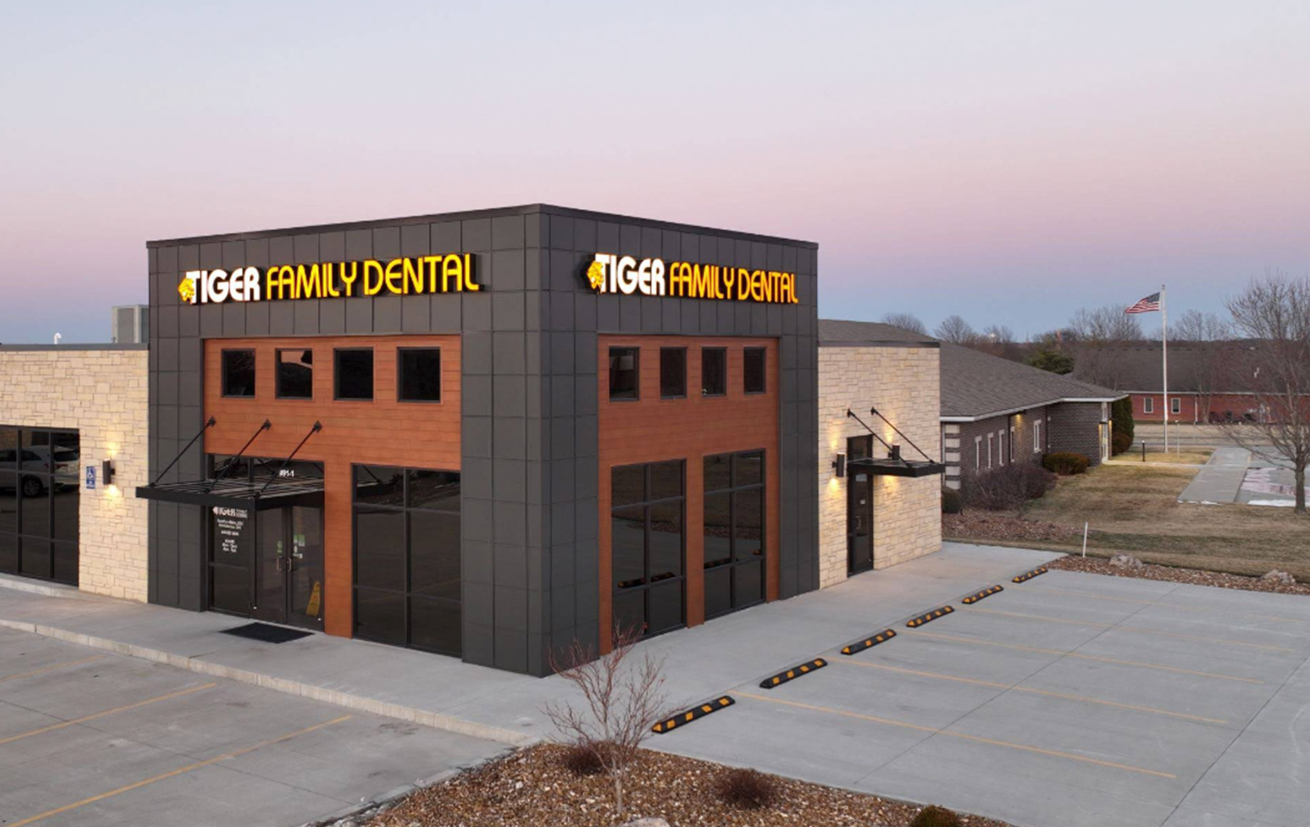 The image shows a modern dental clinic with a prominent sign reading  TIGER FAMILY DENTAL  on a building with a black roof, set against a clear sky at dusk.