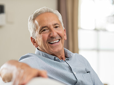 An elderly man with white hair, wearing a blue shirt, sitting on a couch with a smile.