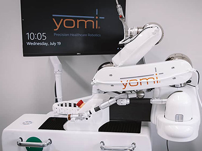 The image shows a robotic arm positioned next to a sign with the word  YOMI  on it, indicating the presence of a healthcare robot within a medical setting.