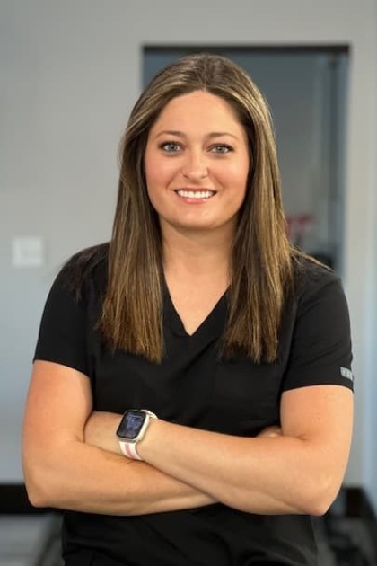 The image shows a woman standing with her arms crossed, wearing a black shirt and a name tag, against a blurred background that appears to be an indoor setting with exercise equipment.