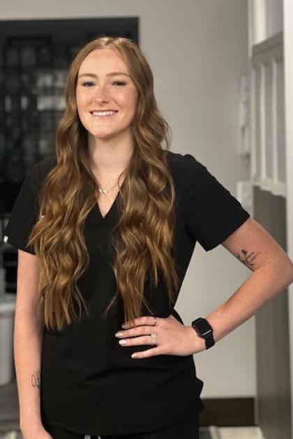 The image shows a woman wearing a black scrub top, standing with her hands on her hips, smiling at the camera. She has long hair and appears to be posing for the photograph.