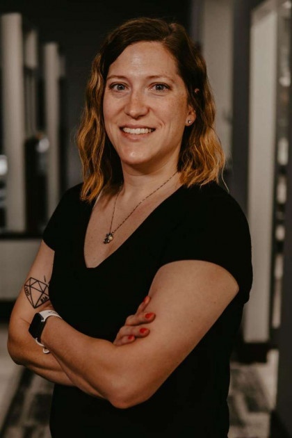 The image features a woman standing with her arms crossed, smiling at the camera, wearing a black shirt and glasses.
