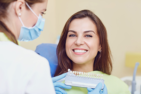 The image shows a dental professional with a smiling expression holding up a dental tool, likely during a dental appointment, with a patient seated in front of them.