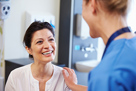 The image shows two individuals in what appears to be a medical setting one person is seated and smiling while another stands behind them, both are engaged in a conversation, with visible medical equipment and a professional environment suggesting a healthcare context.