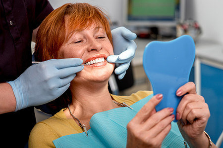 A woman with red hair sitting in a dental chair, holding a blue dental model, smiling at the camera while a dentist works on her teeth.