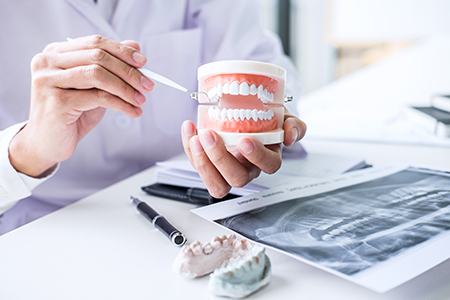 Image 1  A dental professional holding a tray with a model set of teeth and viewing it through a magnifying glass, with a background showing medical equipment and a skeletal model.  Image 2  The same dental professional holding the same model set of teeth in front of a display of dental implants and tools, with an X-ray image visible in the background.