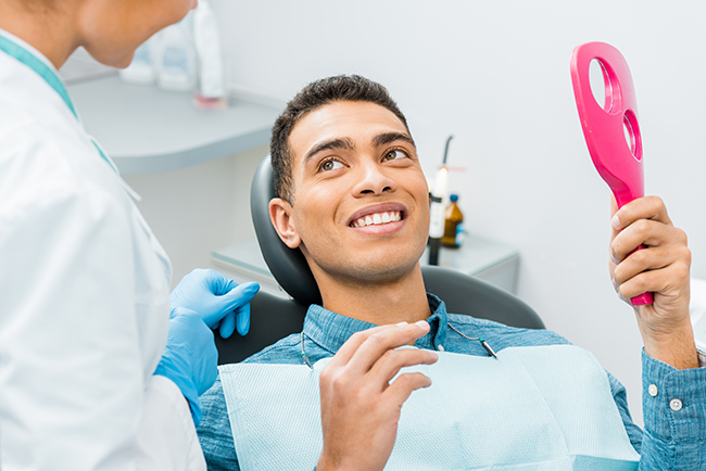 The image shows a man sitting in a dental chair with a smile on his face, holding a pink object that appears to be an oral hygiene tool, while being attended to by a dentist who is smiling and wearing a mask.