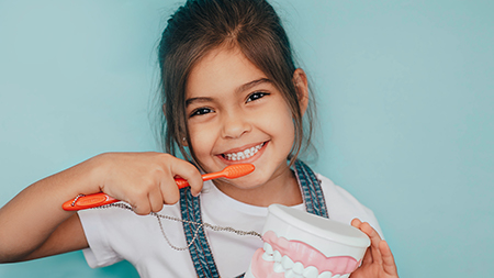 The image features a young girl with a toothbrush, smiling at the camera while brushing her teeth.