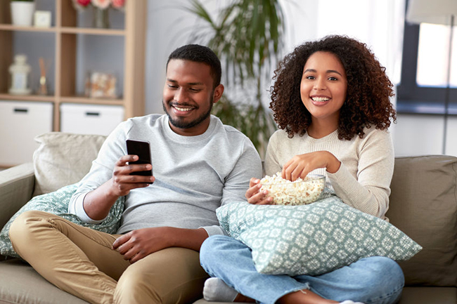 A man and woman sitting on a couch, smiling at each other while holding snacks, watching something on a phone.