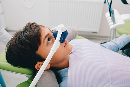 A young man wearing a dental mask with a transparent visor, seated in a dentist s chair during an examination, with his head tilted towards the camera.